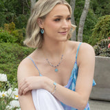 A woman with short blonde hair sits outdoors among greenery, wearing a blue floral dress and vintage-inspired jewelry, including matching blue earrings, a necklace, and the Elena Beaded Bracelet Sky Blue by Holly Yashi. She looks relaxed and gentle.