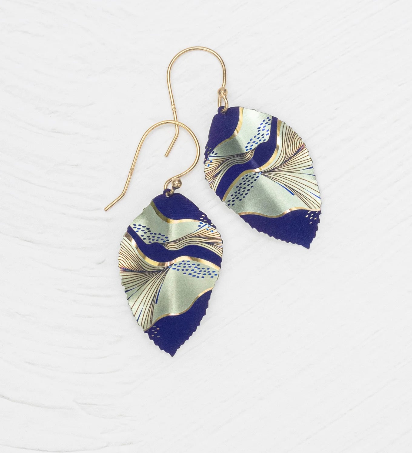 The Eccentric Elm Indigo Earrings by Holly Yashi feature gold hooks and handcrafted leaf pendants with an abstract wavy design in navy, gold, and pale green on a textured white background.
