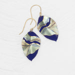 The Eccentric Elm Indigo Earrings by Holly Yashi feature gold hooks and handcrafted leaf pendants with an abstract wavy design in navy, gold, and pale green on a textured white background.