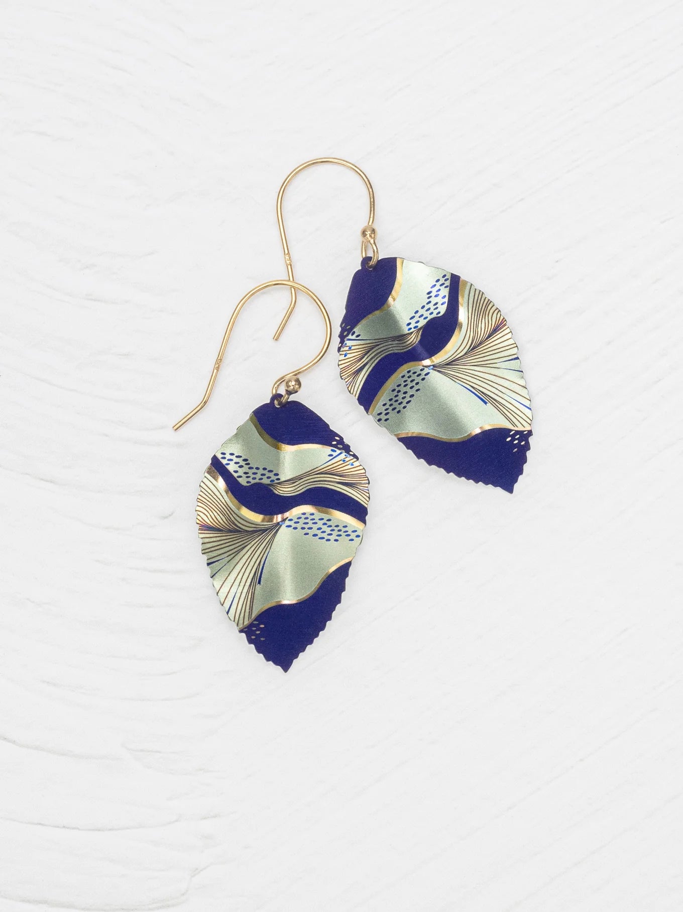 The Eccentric Elm Indigo Earrings by Holly Yashi feature gold hooks and handcrafted leaf pendants with an abstract wavy design in navy, gold, and pale green on a textured white background.