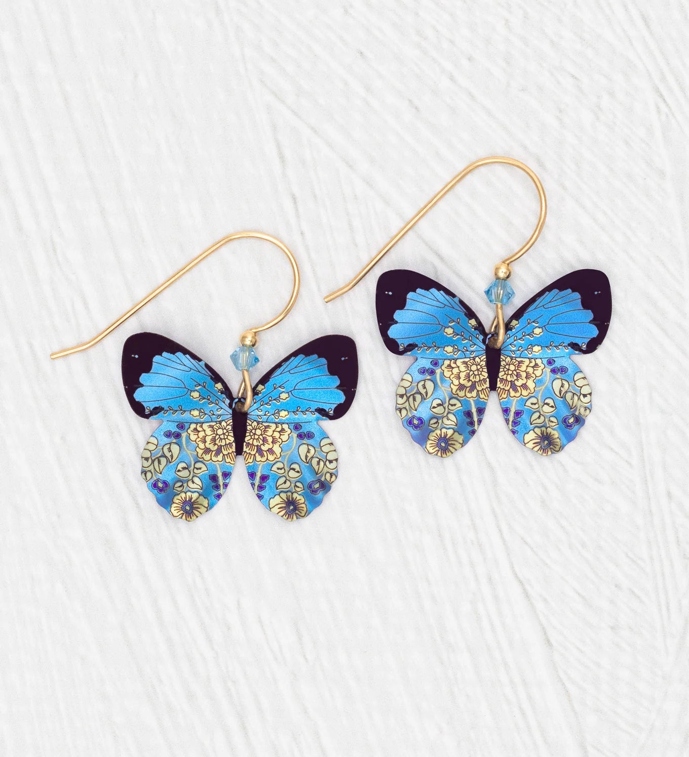 The Holly Yashi Blooming Butterfly Blue Radiance Earrings feature butterfly shapes with vibrant blue wings, intricate gold and yellow floral patterns, and gold-filled wires, shown against a white textured background.