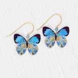 The Holly Yashi Blooming Butterfly Blue Radiance Earrings feature butterfly shapes with vibrant blue wings, intricate gold and yellow floral patterns, and gold-filled wires, shown against a white textured background.