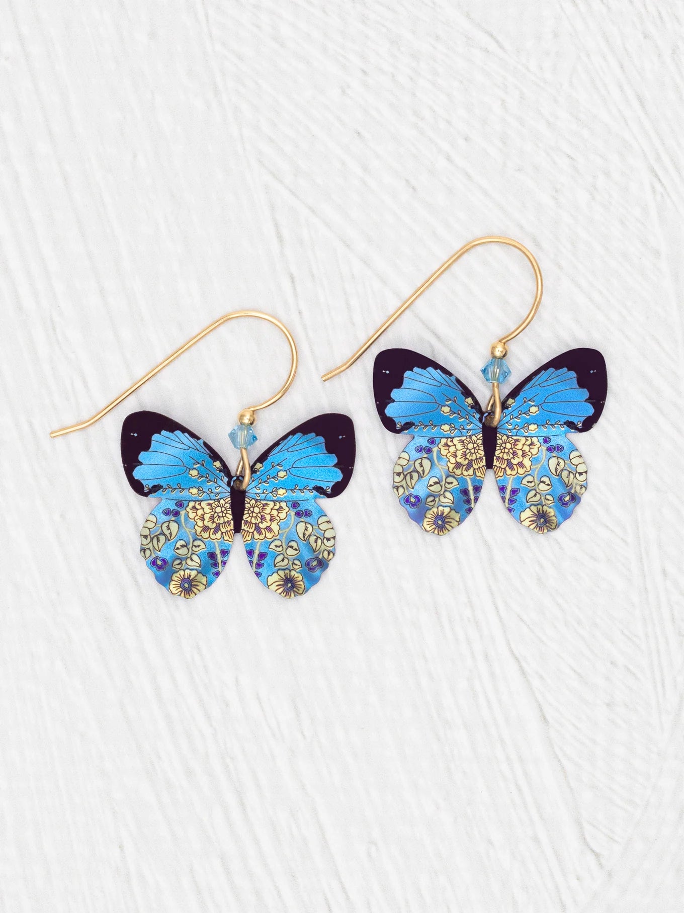 The Holly Yashi Blooming Butterfly Blue Radiance Earrings feature butterfly shapes with vibrant blue wings, intricate gold and yellow floral patterns, and gold-filled wires, shown against a white textured background.