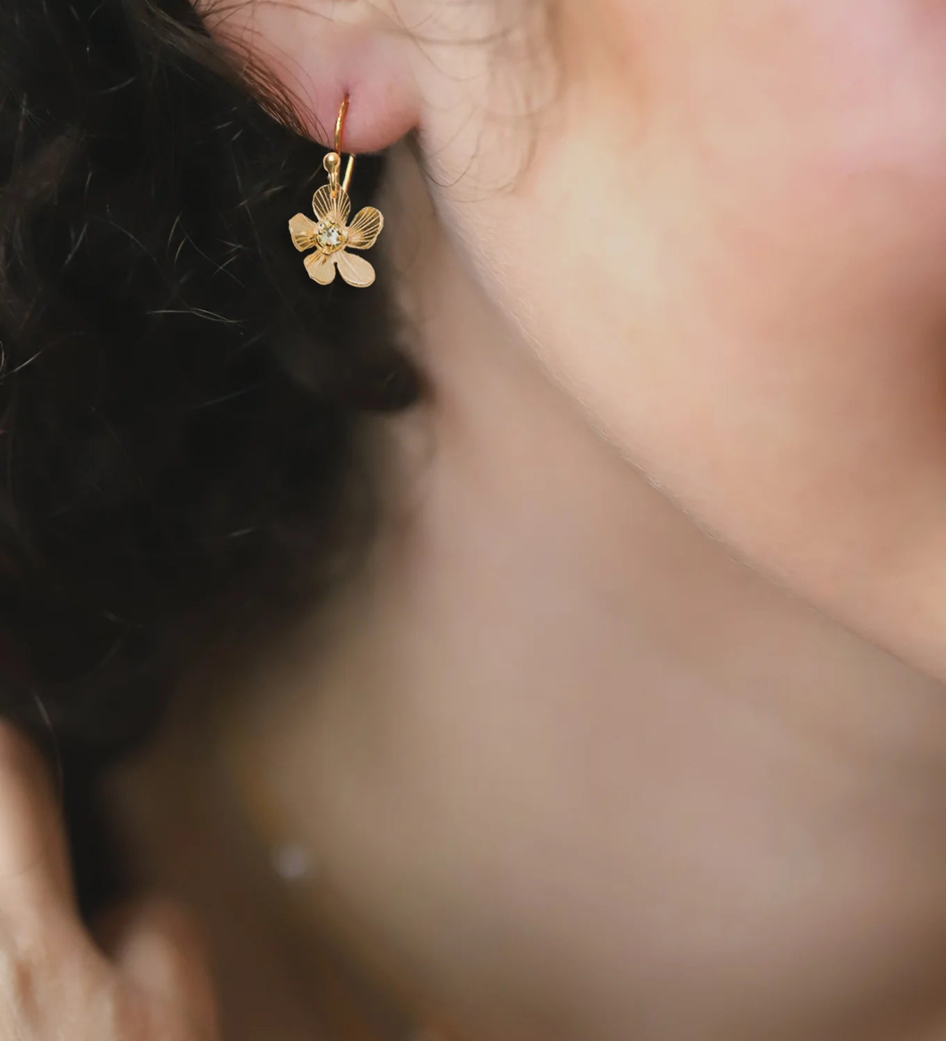 Close-up of a womans ear wearing Holly Yashis Plumeria Drop Gold Earrings. The blurred background highlights her curly hair and smooth skin, showcasing the intricate detail of this feminine jewelry from Holly Yashi.