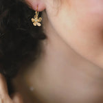 Close-up of a womans ear wearing Holly Yashis Plumeria Drop Gold Earrings. The blurred background highlights her curly hair and smooth skin, showcasing the intricate detail of this feminine jewelry from Holly Yashi.