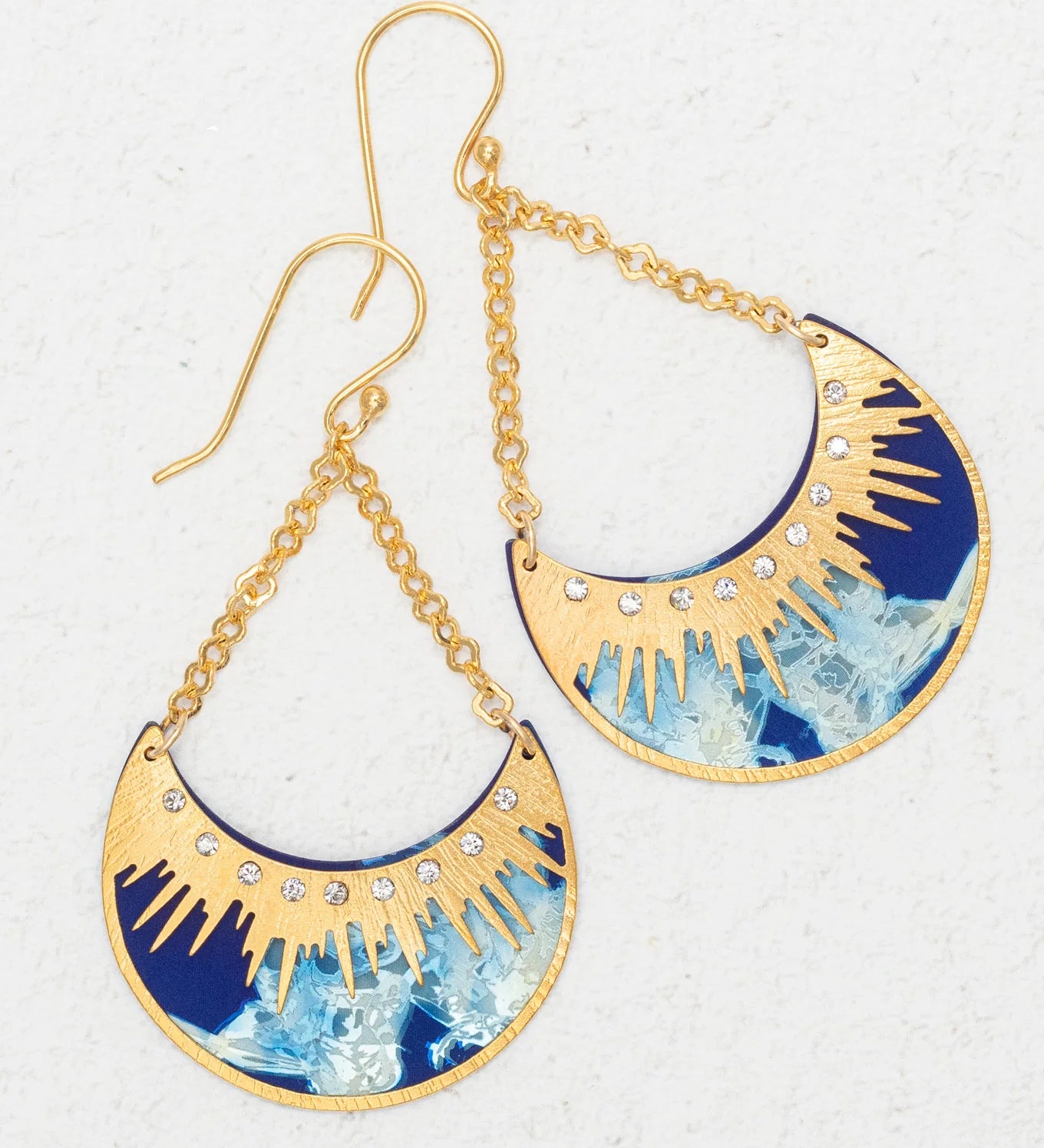 The Selena/ Midnight Blue earrings by Holly Yashi feature gold and blue crescent shapes with sunburst designs, small clear gemstones, and dangling gold chain hooks. Hypoallergenic and shown on a textured white surface.