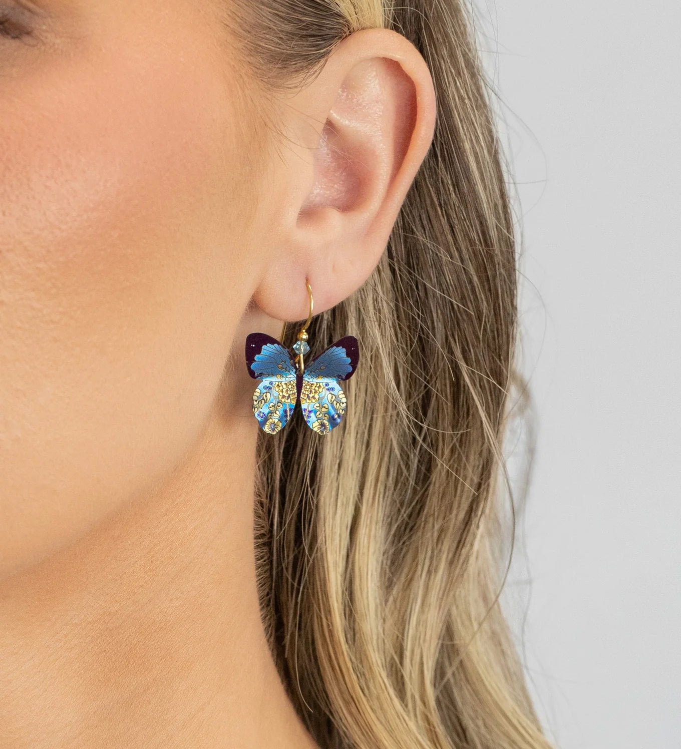A close-up shows a woman with blonde hair partially tucked behind her ear, wearing Holly Yashi’s ER: Blooming Butterfly Radiant Purple earrings. These feature hand-dipped niobium and gold-filled wires with blue, purple, and gold crystal accents.