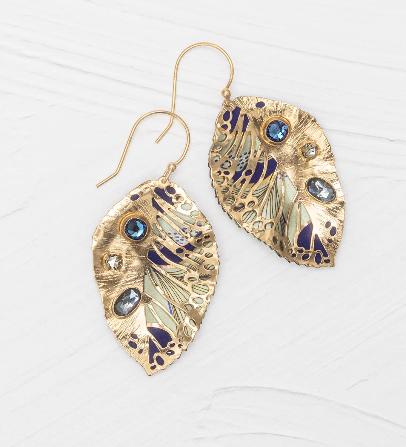 The Holly Yashi Perennial Elm Indigo Earrings are gold, leaf-shaped with blue, silver, and purple accents, featuring blue and clear gemstones. Displayed on a white textured surface.