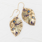 The Holly Yashi Perennial Elm Indigo Earrings are gold, leaf-shaped with blue, silver, and purple accents, featuring blue and clear gemstones. Displayed on a white textured surface.