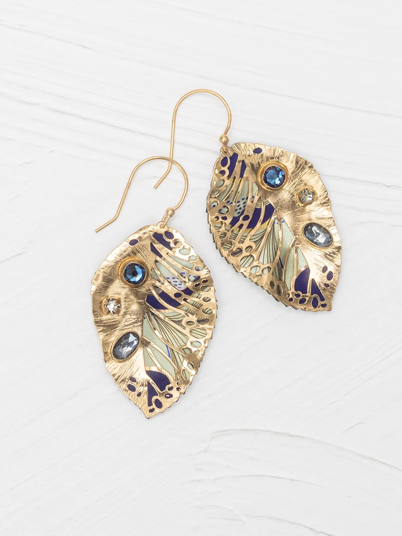 The Holly Yashi Perennial Elm Indigo Earrings are gold, leaf-shaped with blue, silver, and purple accents, featuring blue and clear gemstones. Displayed on a white textured surface.
