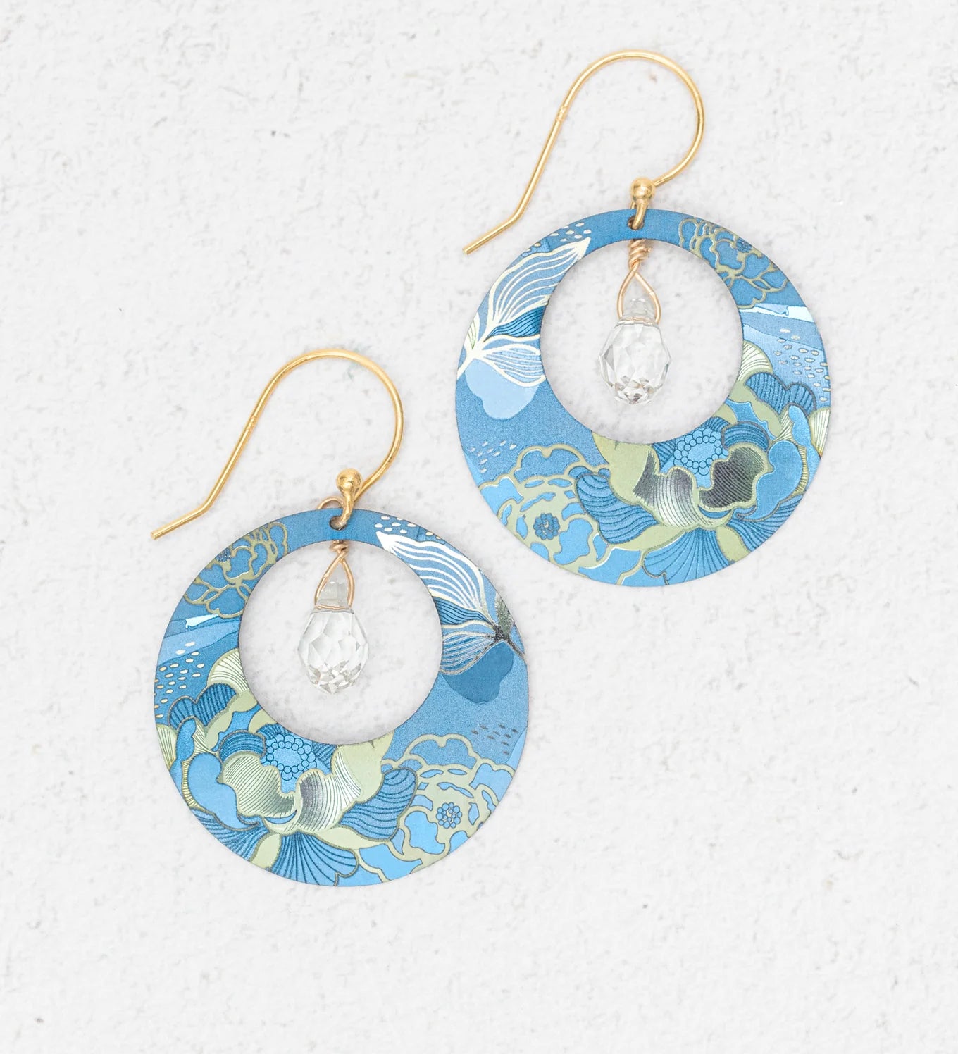The Claire Sky Blue Earrings by Holly Yashi feature handcrafted round frames with blue, green, and gold floral patterns, gold hooks, and a clear faceted bead at the center—perfect for any occasion.