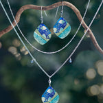 A pair of earrings and two necklaces, including the Holly Yashi Lani Kai Blue Necklace with matching blue, green, and gold pendants, hang from a curved branch against a softly lit blurred natural background. Hypoallergenic jewelry.