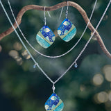 A pair of earrings and two necklaces, including the Holly Yashi Lani Kai Blue Necklace with matching blue, green, and gold pendants, hang from a curved branch against a softly lit blurred natural background. Hypoallergenic jewelry.
