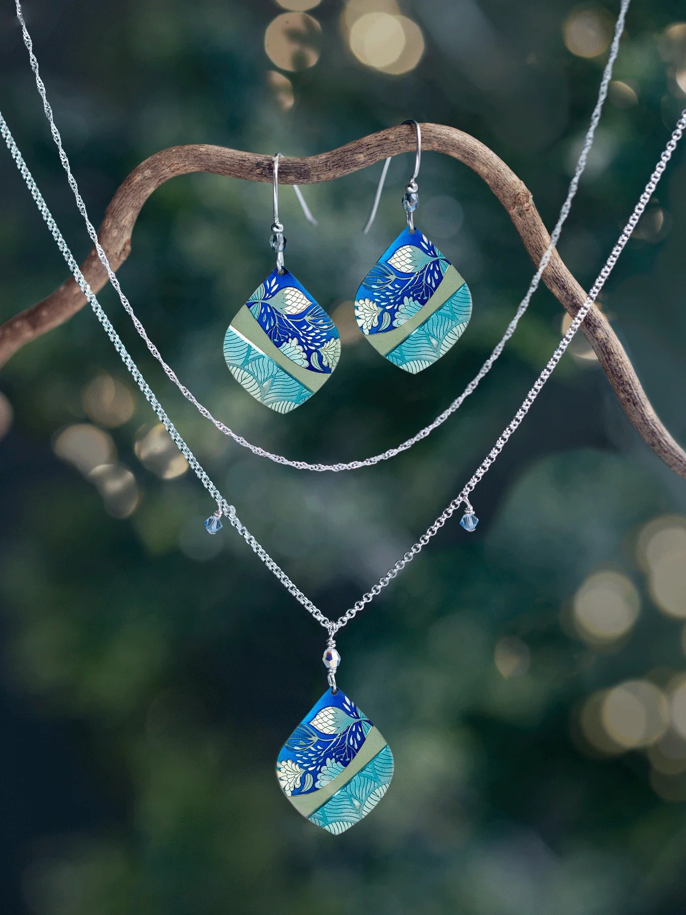 A pair of earrings and two necklaces, including the Holly Yashi Lani Kai Blue Necklace with matching blue, green, and gold pendants, hang from a curved branch against a softly lit blurred natural background. Hypoallergenic jewelry.