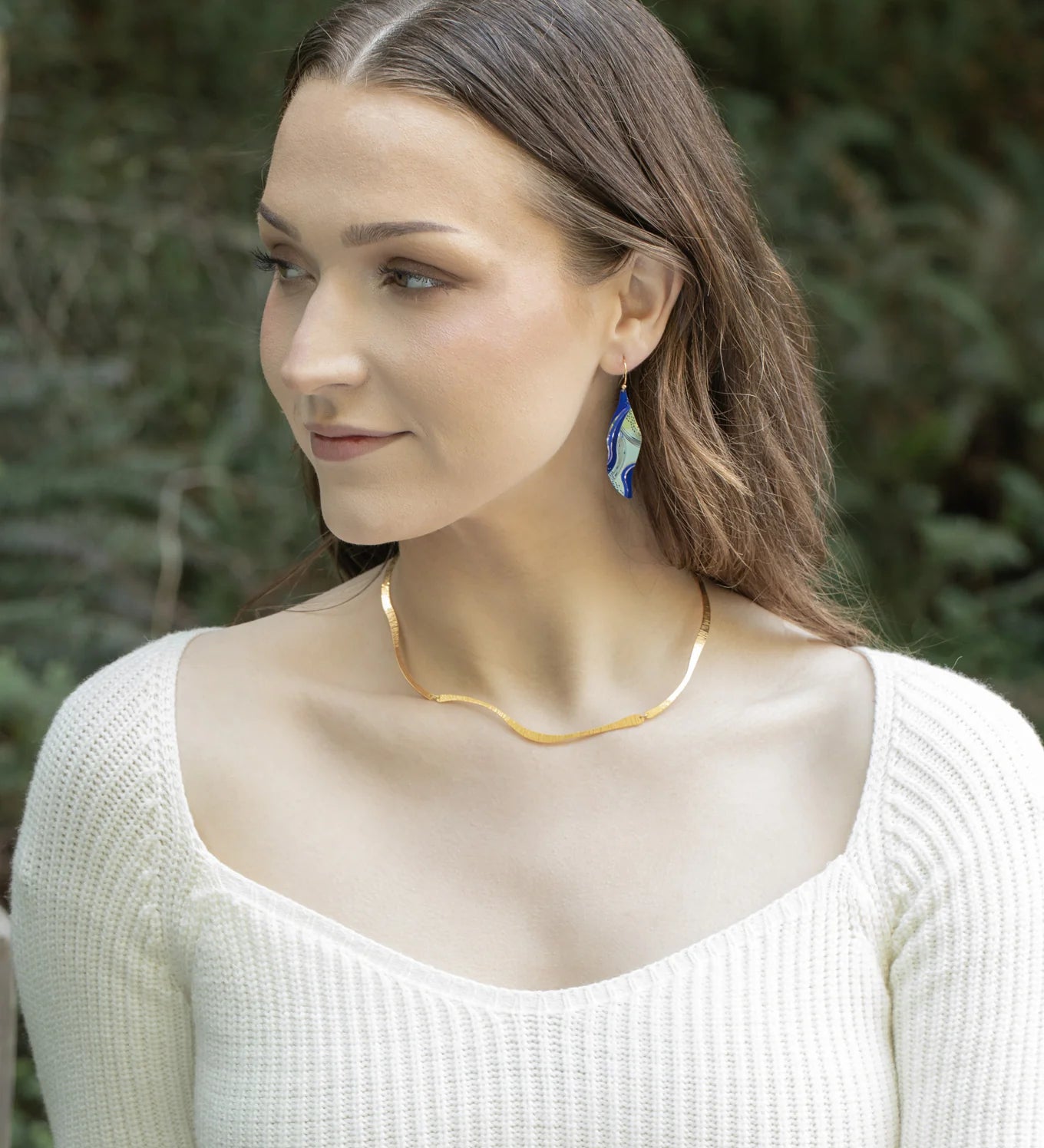 A woman with long brown hair wears a cream ribbed top, a wavy gold necklace, and Holly Yashi Turning Leaf Indigo Earrings, outdoors with greenery. She looks to her left and smiles slightly.