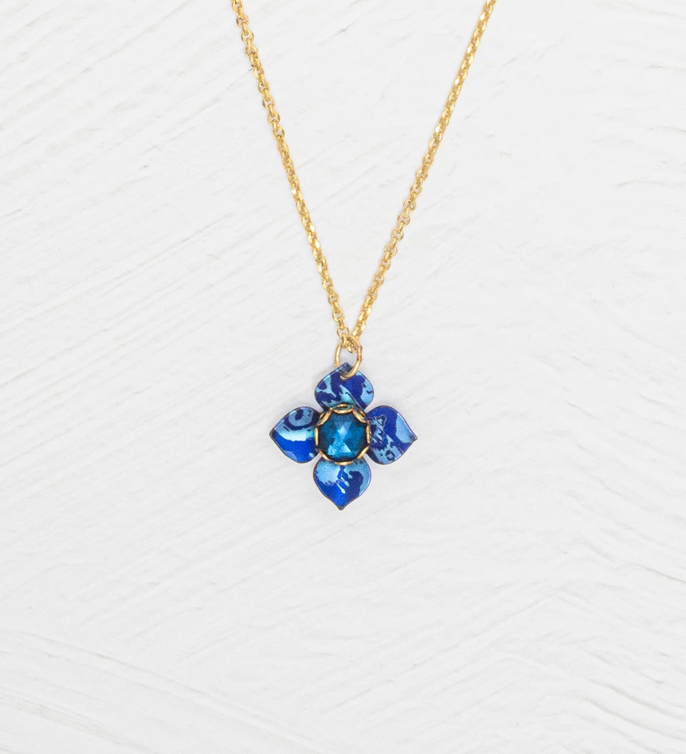 The Petite Fleur Sapphire Blue Necklace by Holly Yashi features a gold chain and a blue flower-shaped pendant with an Austrian crystal, displayed on a white textured background.