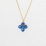 The Petite Fleur Sapphire Blue Necklace by Holly Yashi features a gold chain and a blue flower-shaped pendant with an Austrian crystal, displayed on a white textured background.
