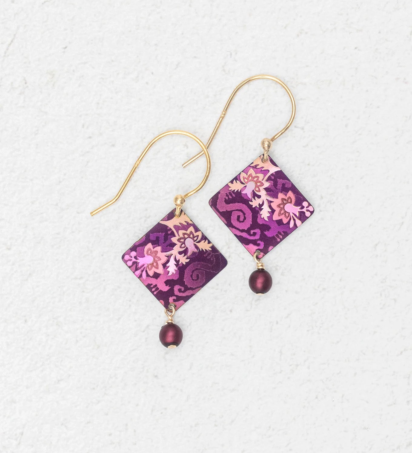 A pair of Holly Yashi Garden Soiree Vintage Burgundy Earrings, featuring gold hooks and diamond-shaped purple and pink patterned charms with a dangling round purple bead, are displayed on a light textured background.