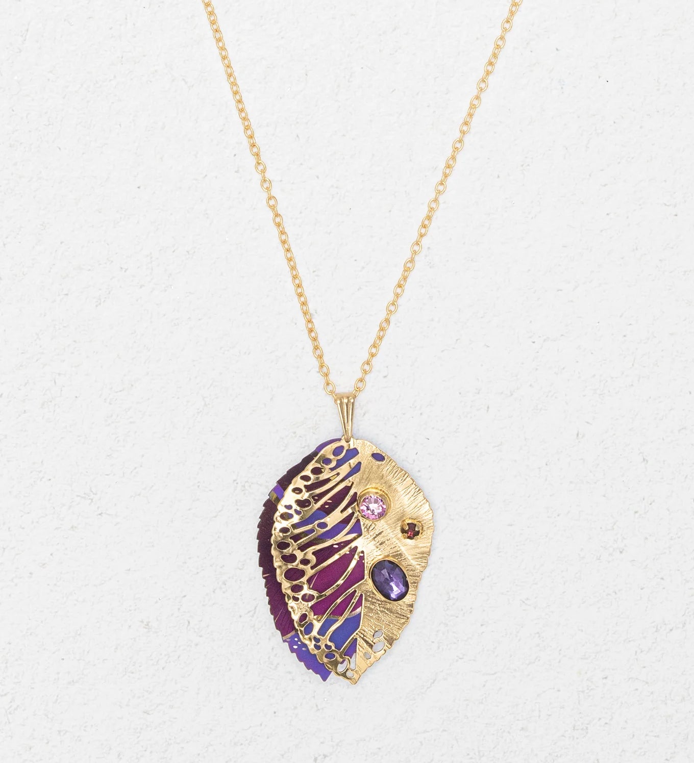 The Perennial Elm Purple Necklace by Holly Yashi features a gold pendant shaped like a stylized leaf with intricate cut-outs and purple, pink, and red gemstones—a striking accessory for any occasion. Displayed on a light textured background.