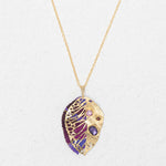 The Perennial Elm Purple Necklace by Holly Yashi features a gold pendant shaped like a stylized leaf with intricate cut-outs and purple, pink, and red gemstones—a striking accessory for any occasion. Displayed on a light textured background.