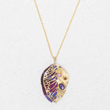 The Perennial Elm Purple Necklace by Holly Yashi features a gold pendant shaped like a stylized leaf with intricate cut-outs and purple, pink, and red gemstones—a striking accessory for any occasion. Displayed on a light textured background.