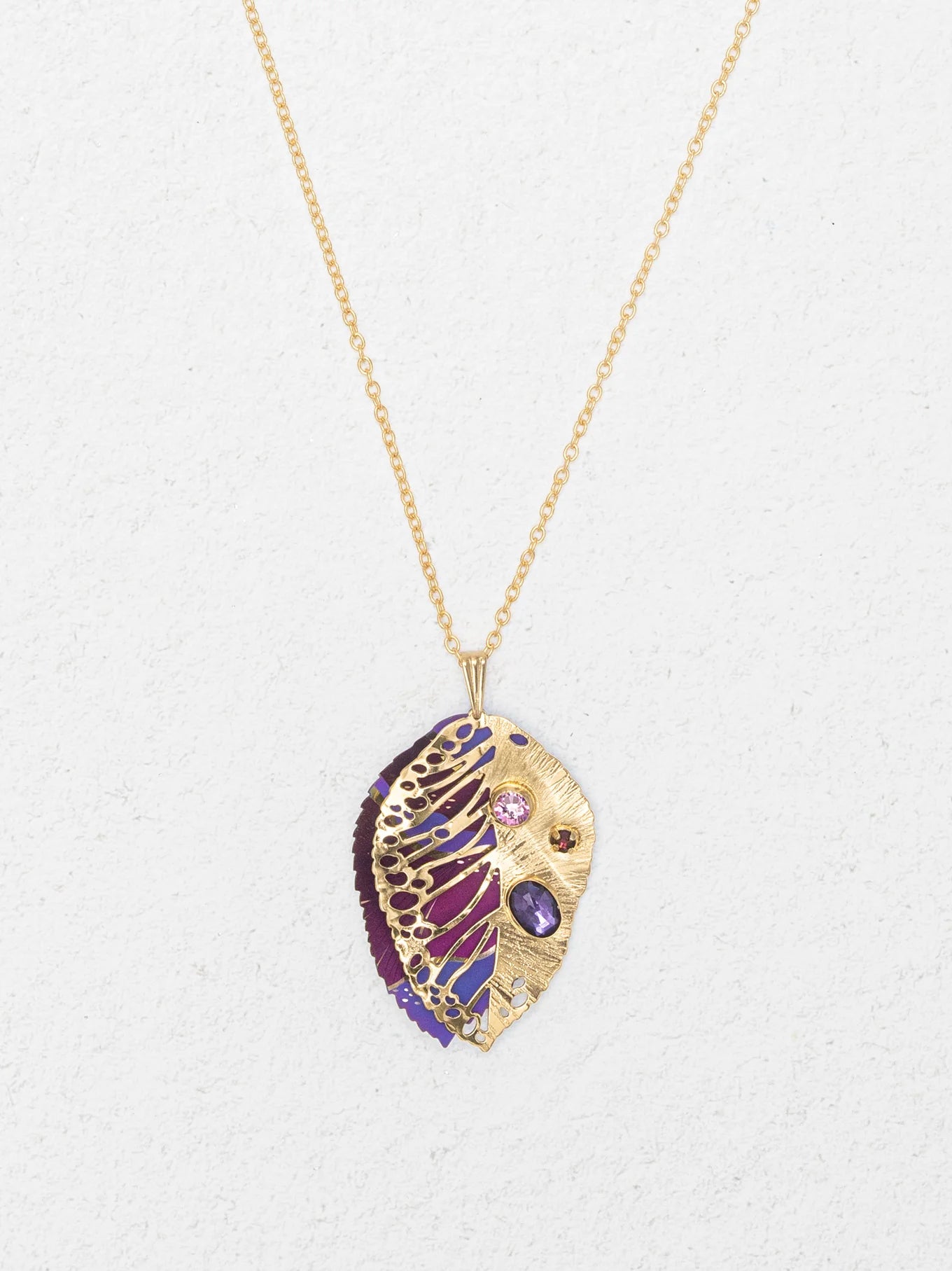 The Perennial Elm Purple Necklace by Holly Yashi features a gold pendant shaped like a stylized leaf with intricate cut-outs and purple, pink, and red gemstones—a striking accessory for any occasion. Displayed on a light textured background.