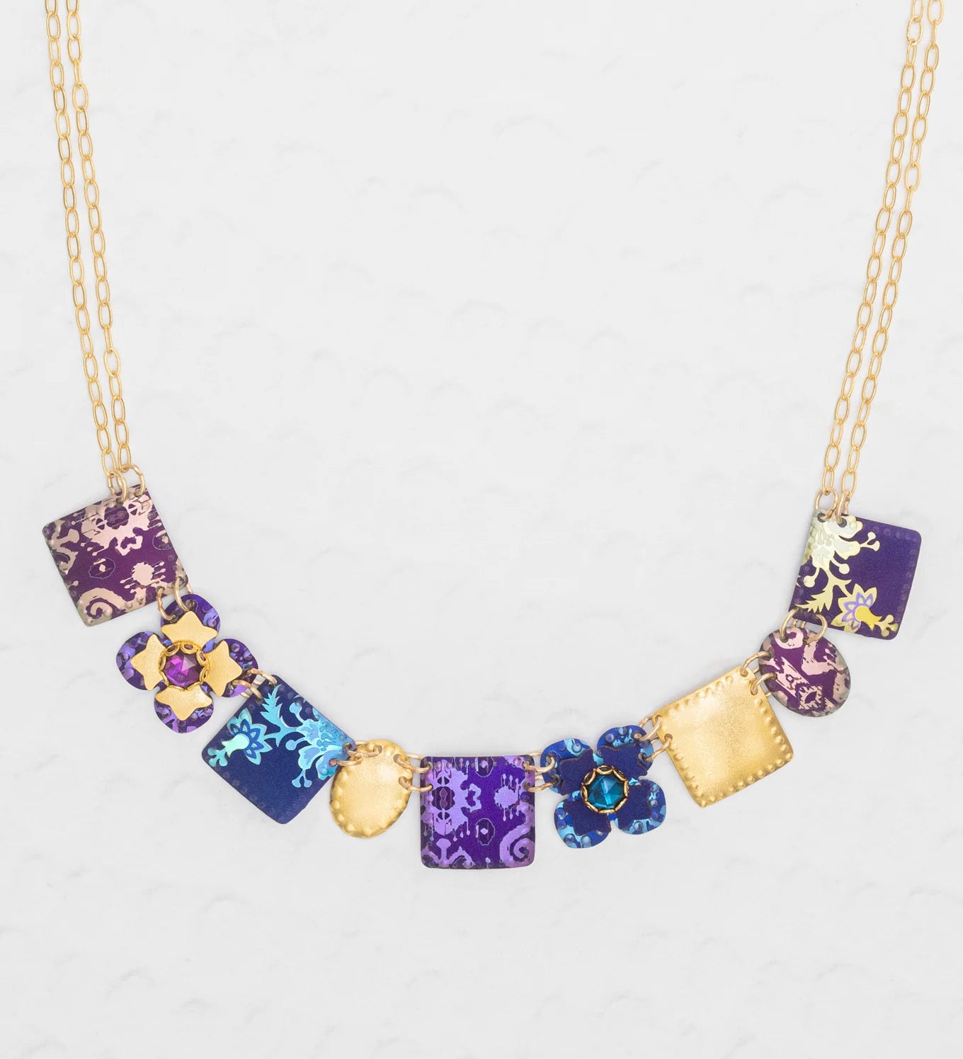 The Eliza Tiled Multi-Color Necklace by Holly Yashi features square and flower-shaped pendants in purple, blue, and gold with floral patterns and textured designs—an elegant necklace displayed on a light background.