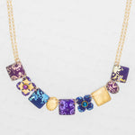 The Eliza Tiled Multi-Color Necklace by Holly Yashi features square and flower-shaped pendants in purple, blue, and gold with floral patterns and textured designs—an elegant necklace displayed on a light background.