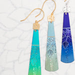 The Midnight Blue Tula Earrings by Holly Yashi feature three vibrant floral designs on an ombré base: turquoise to blue, light blue to gray, and dark blue to purple. Made with 18k gold-filled and silver hooks, these hypo-allergenic earrings are set against a crisp white background.