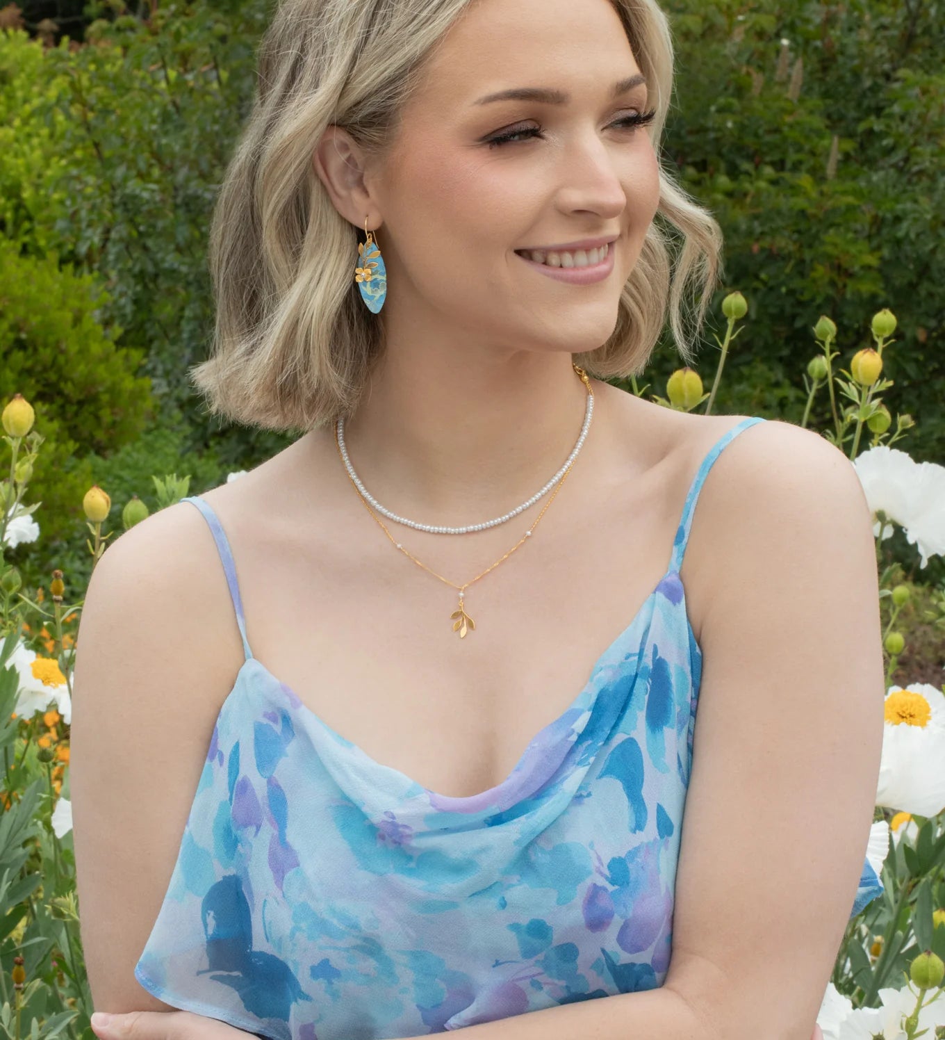 A woman with blonde wavy hair smiles in a garden of white flowers and greenery, wearing Holly Yashi’s Alyssa Sky Blue Earrings and a light blue floral dress.