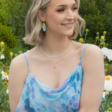 A woman with blonde wavy hair smiles in a garden of white flowers and greenery, wearing Holly Yashi’s Alyssa Sky Blue Earrings and a light blue floral dress.