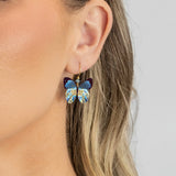 Close-up of a person wearing Holly Yashi’s Blooming Butterfly Blue Radiance Earrings, featuring glittery blue wings and gold-filled wires. Blonde hair is tucked behind the ear, highlighting these crystal-accented earrings against a gray background.