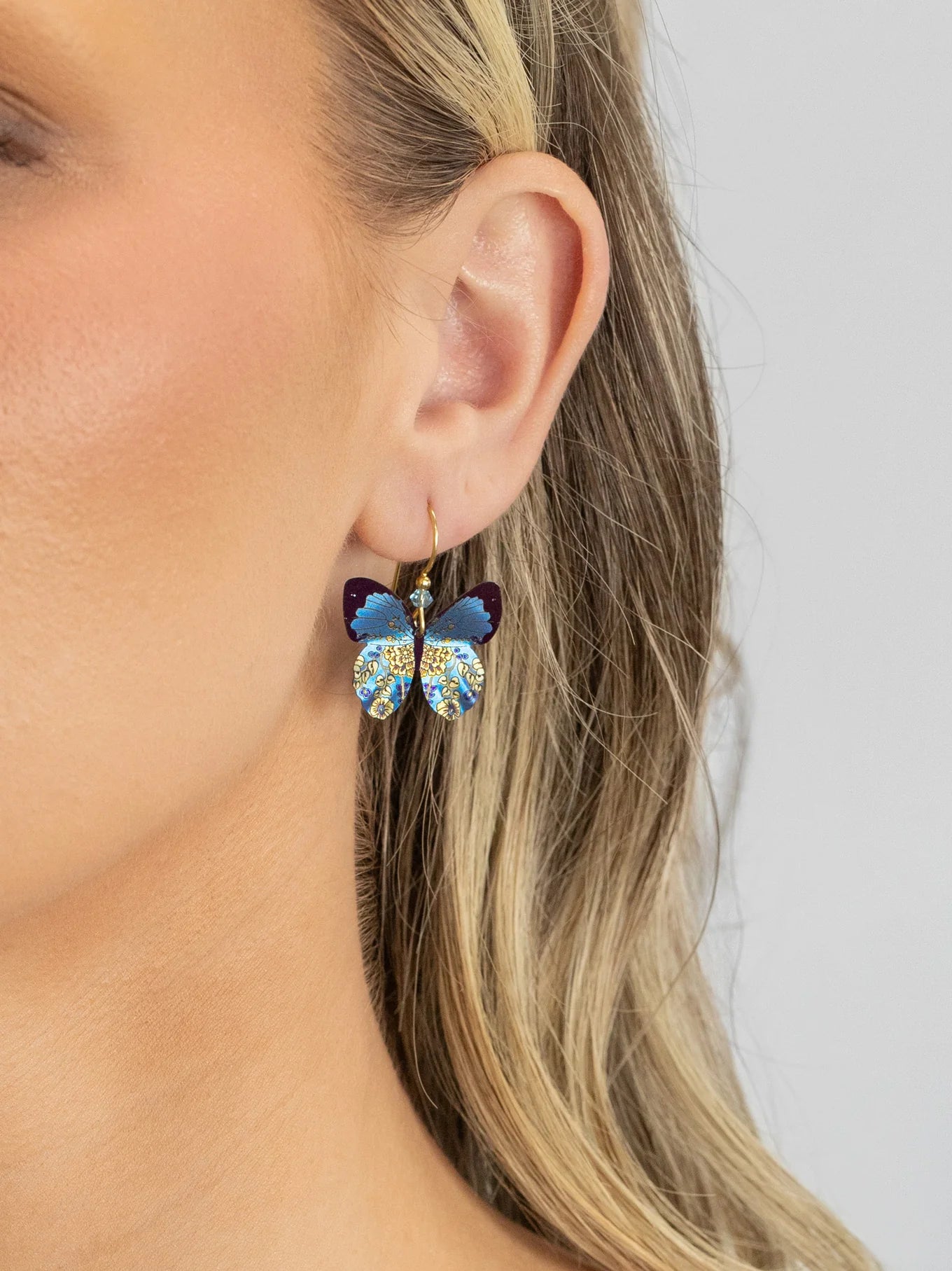 Close-up of a person wearing Holly Yashi’s Blooming Butterfly Blue Radiance Earrings, featuring glittery blue wings and gold-filled wires. Blonde hair is tucked behind the ear, highlighting these crystal-accented earrings against a gray background.
