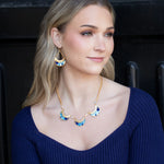 A woman with long blonde hair wears a blue knit top, colorful crescent earrings, and the Selena Necklace-Midnight Blue by Holly Yashi, sitting before a dark background and smiling softly as she looks to the side.