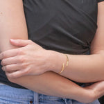 A person in a black short-sleeved top and light blue jeans crosses their arms, wearing a thin gold bracelet and the Holly Yashi Zara Cuff Gold Bracelet on one wrist. Their face is not visible.