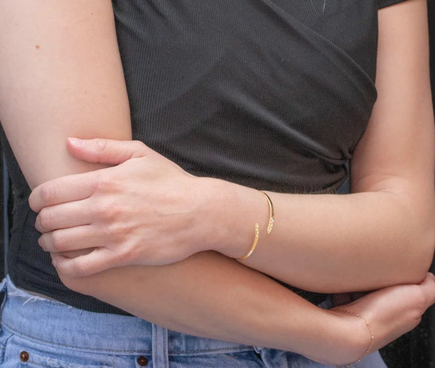 A person in a black short-sleeved top and light blue jeans crosses their arms, wearing a thin gold bracelet and the Holly Yashi Zara Cuff Gold Bracelet on one wrist. Their face is not visible.