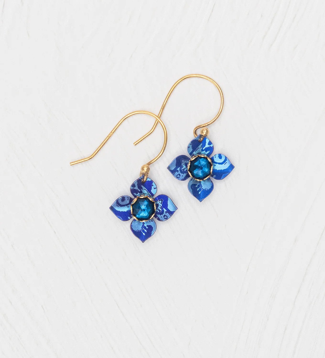 The Petite Fleur Sapphire Blue Earrings by Holly Yashi feature gold hooks and flower-shaped pendants with Austrian crystal blue stones, beautifully displayed on a white textured surface.