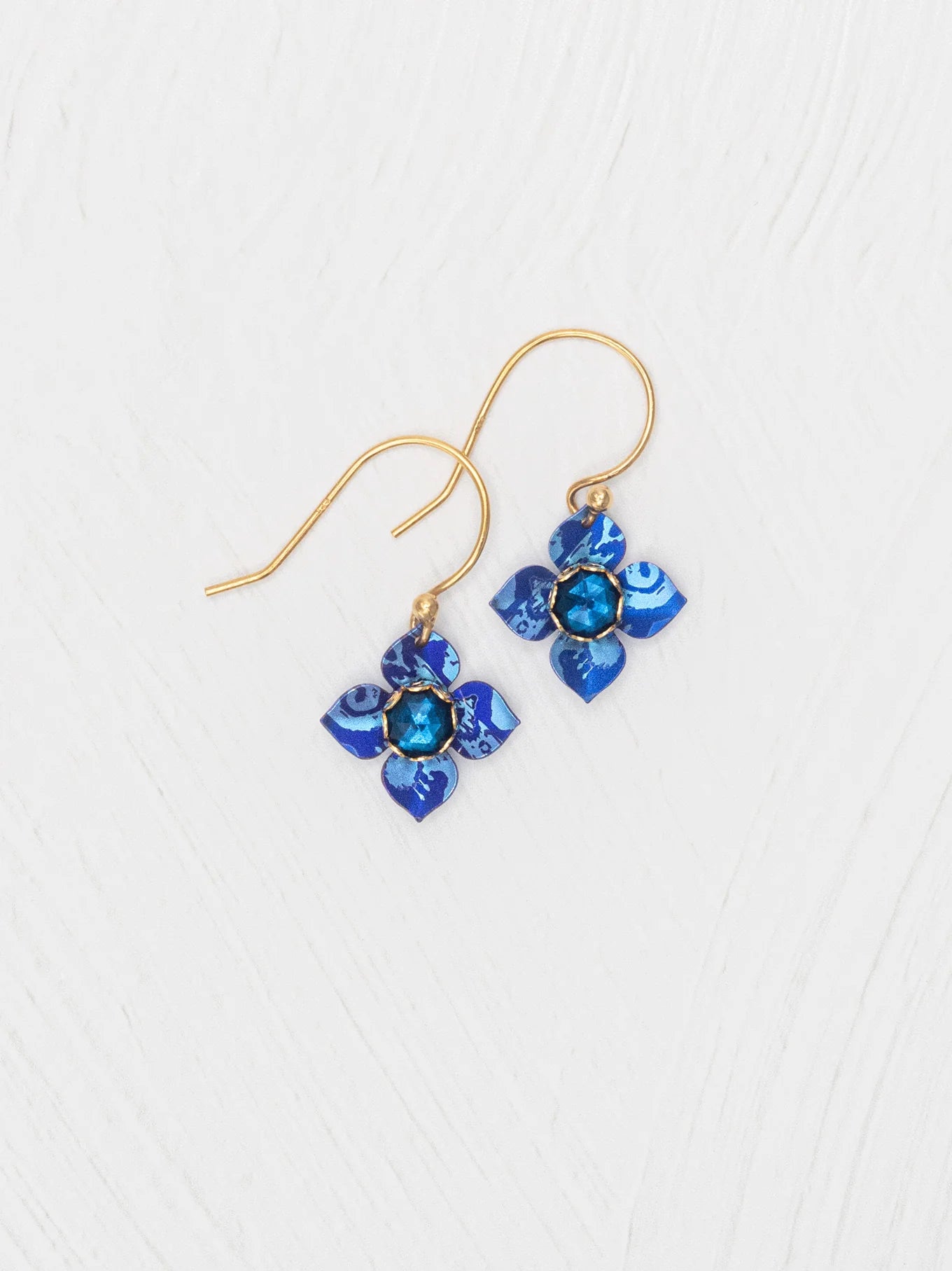 The Petite Fleur Sapphire Blue Earrings by Holly Yashi feature gold hooks and flower-shaped pendants with Austrian crystal blue stones, beautifully displayed on a white textured surface.