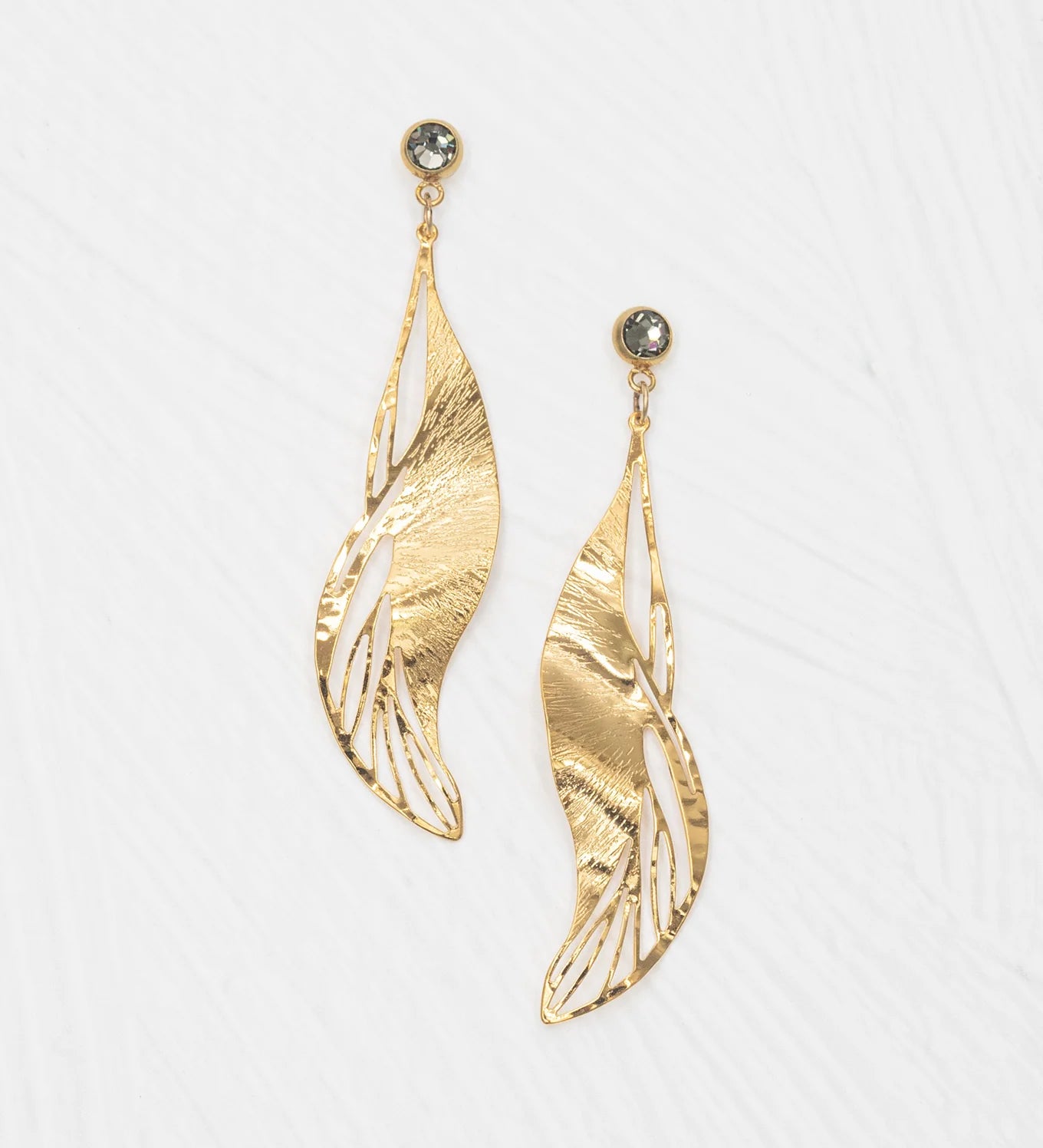 The Holly Yashi ER: Gilded Leaf Post Gold earrings feature gold, leaf-shaped drops with textured, wavy silhouettes and open cut-outs, each accented by a round crystal post. Shown against a white background.