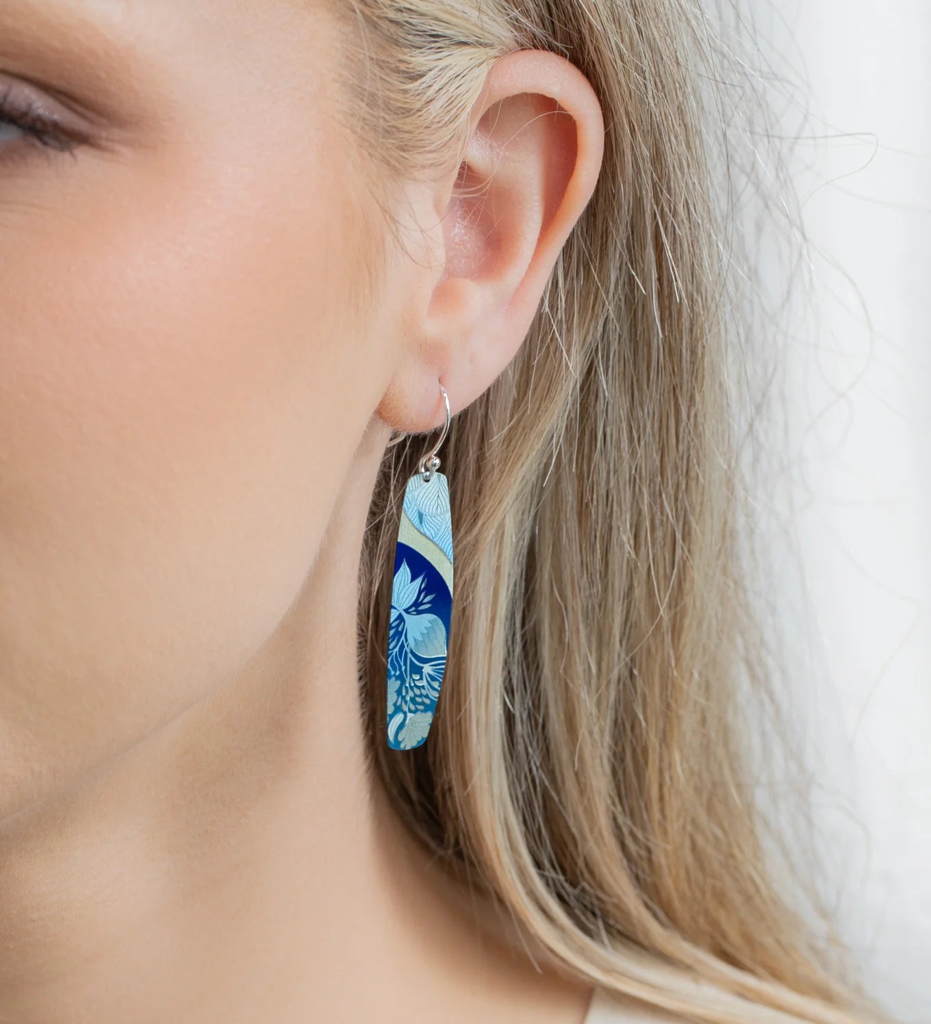 A close-up of a womans ear wearing Holly Yashis Malia Kai Blue Earrings, long oval handcrafted niobium pieces featuring a blue and light green floral pattern. Her blonde hair is tucked behind her ear, showing off the earrings.