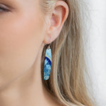 A close-up of a womans ear wearing Holly Yashis Malia Kai Blue Earrings, long oval handcrafted niobium pieces featuring a blue and light green floral pattern. Her blonde hair is tucked behind her ear, showing off the earrings.