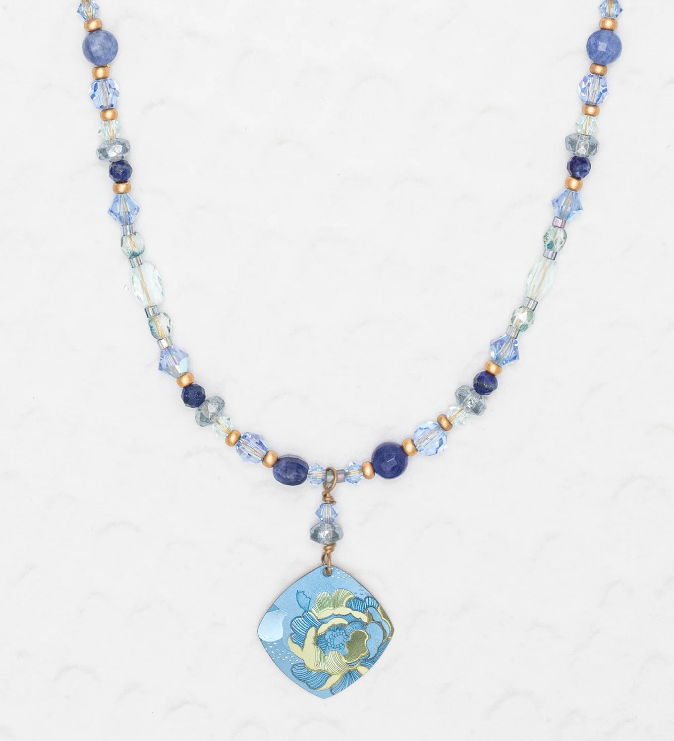 The Holly Yashi Elena Beaded Necklace features sky blue and clear beads with a niobium floral pendant in blue, green, and yellow, shown on a light textured background.