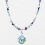 The Holly Yashi Elena Beaded Necklace features sky blue and clear beads with a niobium floral pendant in blue, green, and yellow, shown on a light textured background.