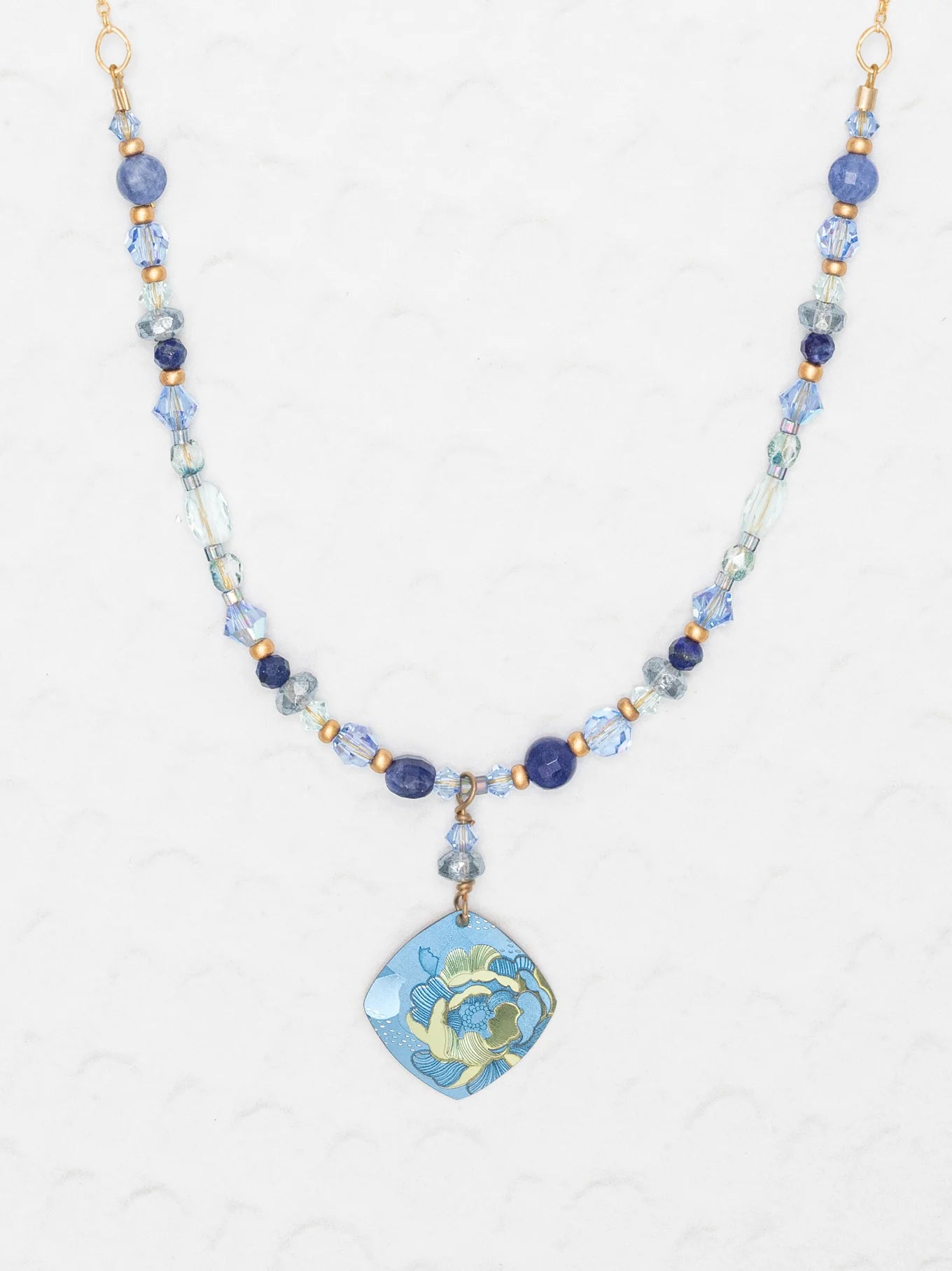 The Holly Yashi Elena Beaded Necklace features sky blue and clear beads with a niobium floral pendant in blue, green, and yellow, shown on a light textured background.