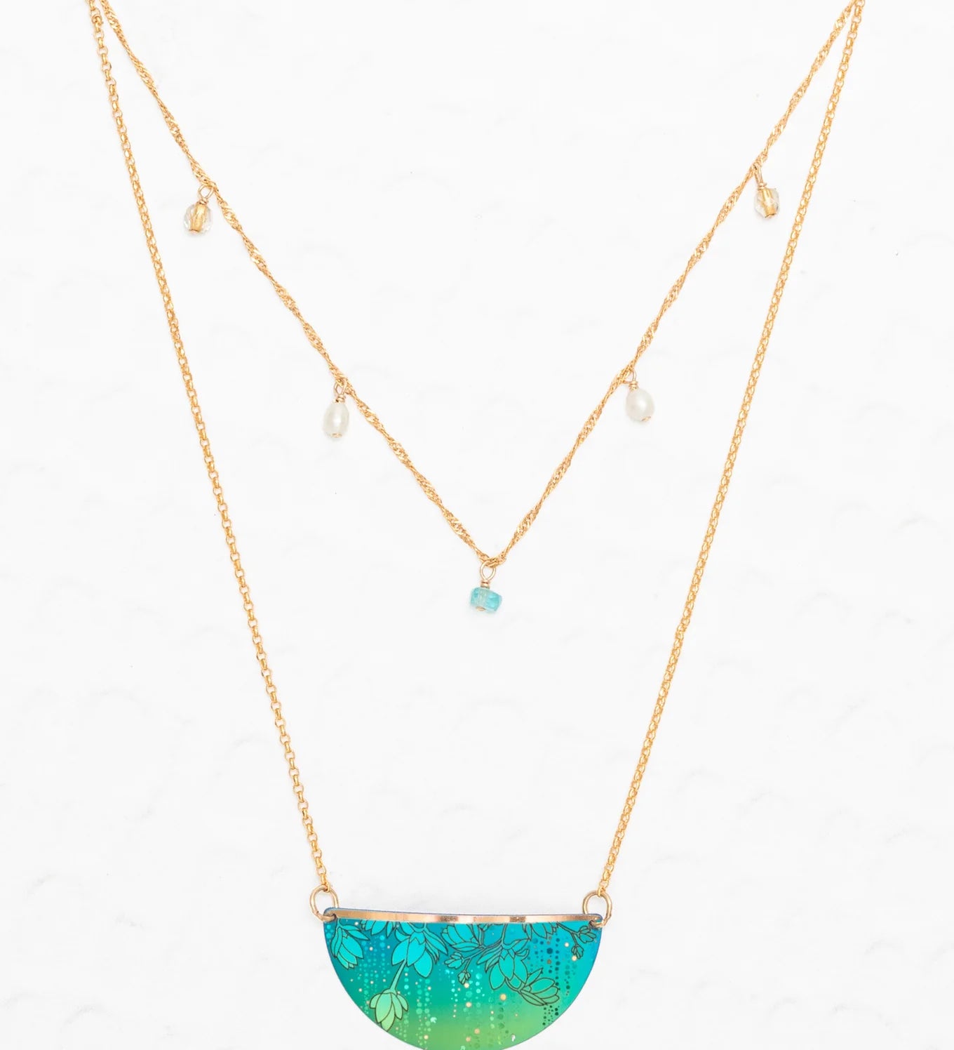 The Magnolia Turquoise/Green Necklace by Holly Yashi features a double-layered gold chain: one with dangling pearls and turquoise bead, and the other with a halfmoon pendant adorned in tropical green and blue florals. Set on a white background.