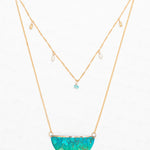 The Magnolia Turquoise/Green Necklace by Holly Yashi features a double-layered gold chain: one with dangling pearls and turquoise bead, and the other with a halfmoon pendant adorned in tropical green and blue florals. Set on a white background.