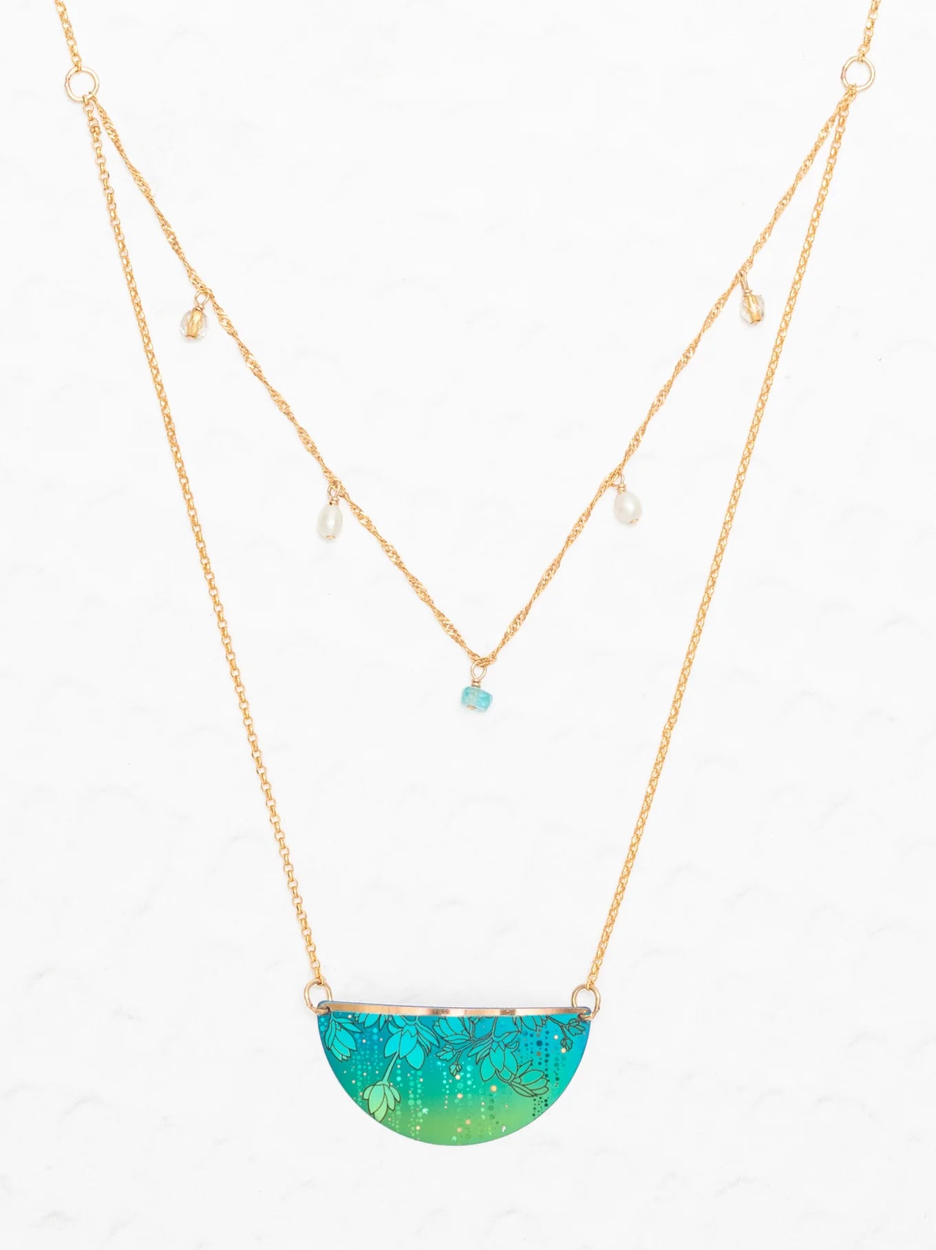 The Magnolia Turquoise/Green Necklace by Holly Yashi features a double-layered gold chain: one with dangling pearls and turquoise bead, and the other with a halfmoon pendant adorned in tropical green and blue florals. Set on a white background.