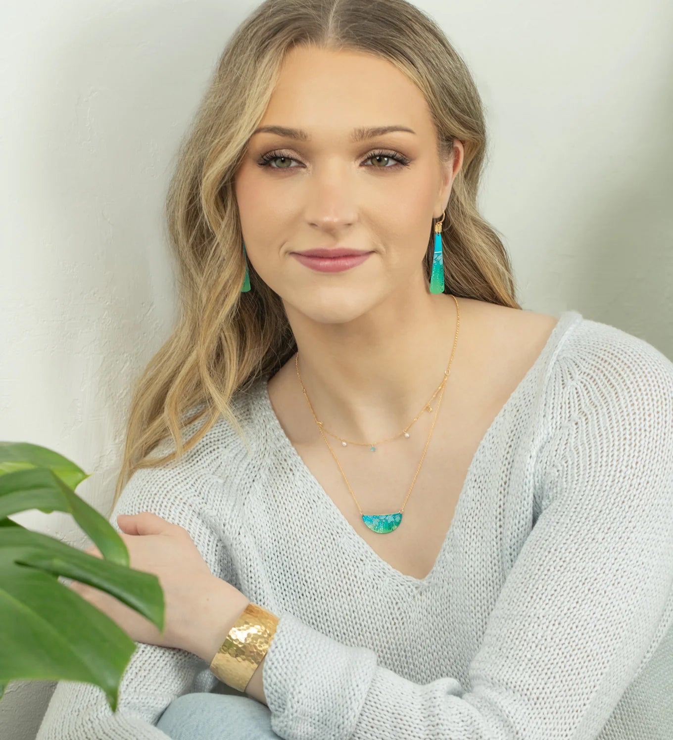 A young woman with long blonde hair wears a light knit sweater, gold jewelry, and the Magnolia Turquoise/Green Necklace by Holly Yashi. She sits beside a green plant, smiling gently at the camera against a light background.