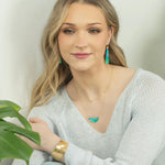 A young woman with long blonde hair wears a light knit sweater, gold jewelry, and the Magnolia Turquoise/Green Necklace by Holly Yashi. She sits beside a green plant, smiling gently at the camera against a light background.