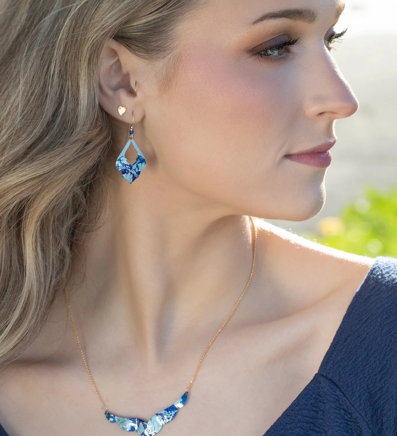 A woman with long blonde hair wearing a blue top and the Holly Yashi Isla Forever Blue Necklace looks to the right in soft natural light.